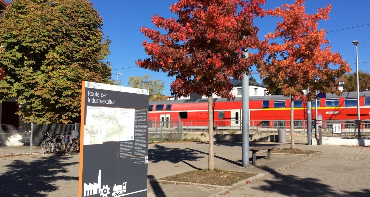 Schild der Route der Industriekultur neben roten Bäumen und einem vorbeifahrenden Zug. Herbstliche Atmosphäre mit klarem Himmel., © Herde Schild der Route der Industriekultur neben roten Bäumen und einem vorbeifahrenden Zug. Herbstliche Atmosphäre mit klarem Himmel., © Herde