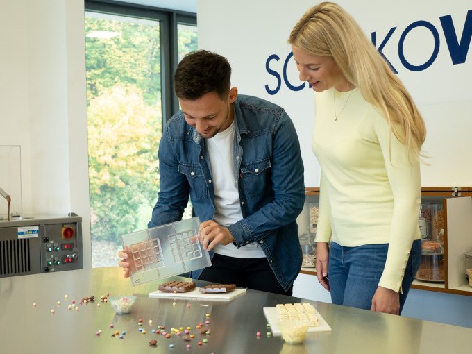 Zwei Personen stehen in einer K&uuml;che und betrachten eine Schokoladenform. Auf dem Tisch liegen bunte Streusel und Schokoladenst&uuml;cke., &copy; Alfred Ritter GmbH &amp; Co. KG | Waldenbuch