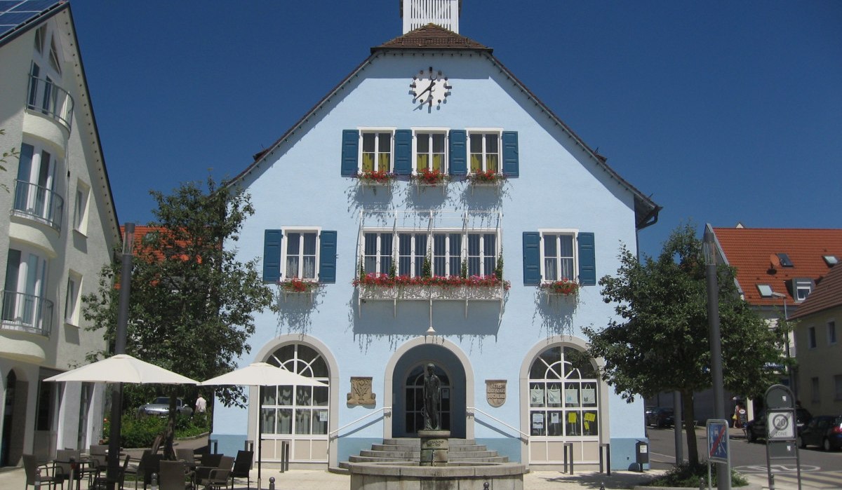 Blaues Gebäude mit Uhr und Blumenkästen, davor ein Brunnen und Café mit Sonnenschirmen, bei sonnigem Wetter., © http://www.rutesheim.de/servlet/PB/menu/1110073/index.html
