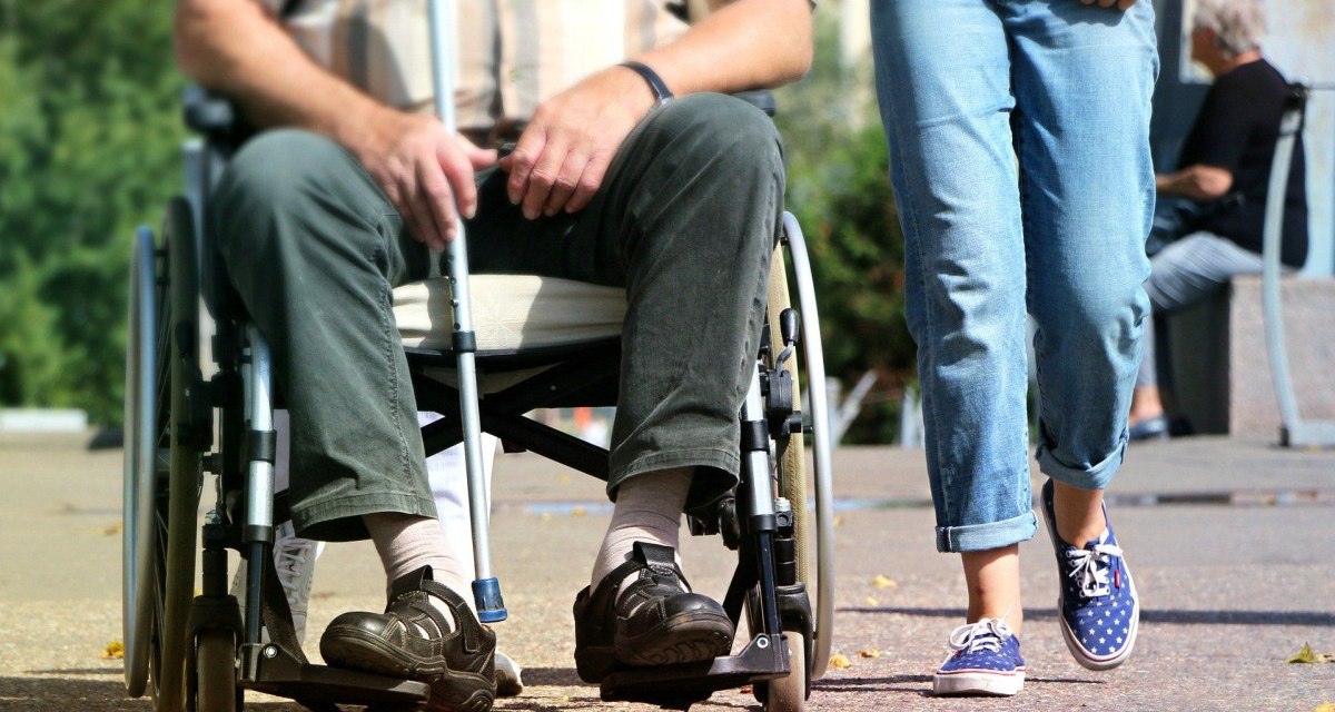 Ein Mann sitzt in einem Rollstuhl auf einem Gehweg, während eine Person in Jeans und Turnschuhen daneben geht., © pixabay Ein Mann sitzt in einem Rollstuhl auf einem Gehweg, während eine Person in Jeans und Turnschuhen daneben geht., © pixabay