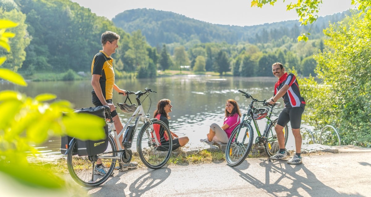 Vier Personen mit Fahrrädern genießen die Natur an einem Seeufer. Zwei sitzen, zwei stehen mit Fahrrädern. Im Hintergrund bewaldete Hügel., © Landratsamt Göppingen Vier Personen mit Fahrrädern genießen die Natur an einem Seeufer. Zwei sitzen, zwei stehen mit Fahrrädern. Im Hintergrund bewaldete Hügel., © Landratsamt Göppingen