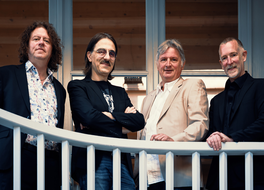 Vier Männer in Anzügen stehen hinter einem weißen Geländer und lächeln in die Kamera. Hintergrund aus Holz und Glas., © Foto Privat Vier Männer in Anzügen stehen hinter einem weißen Geländer und lächeln in die Kamera. Hintergrund aus Holz und Glas., © Foto Privat