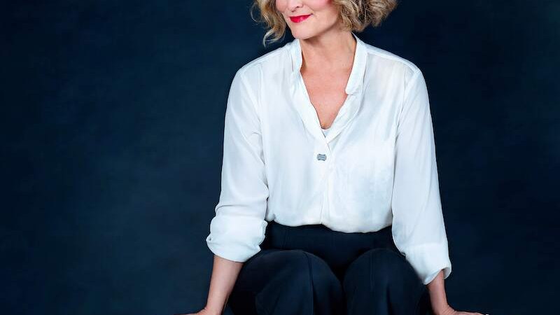 Frau mit blonder Lockenfrisur in weißer Bluse und schwarzer Hose sitzt lächelnd auf einem Tisch vor dunklem Hintergrund., © Mauerwerk GmbH Frau mit blonder Lockenfrisur in weißer Bluse und schwarzer Hose sitzt lächelnd auf einem Tisch vor dunklem Hintergrund., © Mauerwerk GmbH