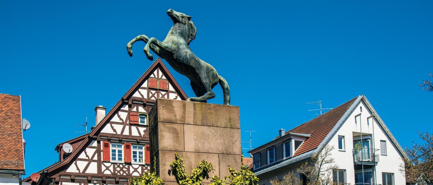 Eine Pferdestatue auf einem Sockel vor traditionellen Fachwerkhäusern in Kirchheim unter Teck, unter klarem, blauem Himmel., © Stuttgart-Marketing GmbH, Sarah Schmid Eine Pferdestatue auf einem Sockel vor traditionellen Fachwerkhäusern in Kirchheim unter Teck, unter klarem, blauem Himmel., © Stuttgart-Marketing GmbH, Sarah Schmid