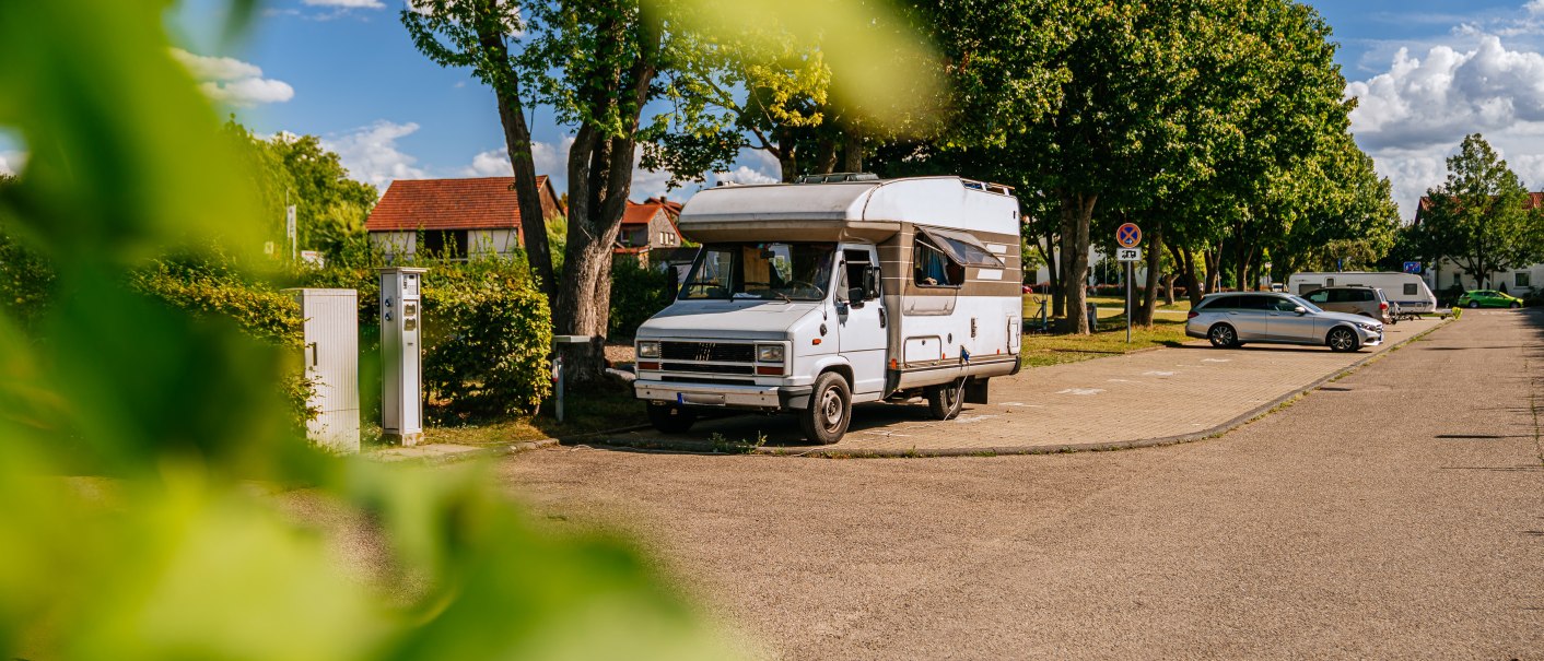Ein Wohnmobil steht auf einem Stellplatz in Marbach am Neckar, umgeben von Bäumen und geparkten Autos. Im Vordergrund sind grüne Blätter zu sehen., © Stuttgart-Marketing GmbH, Thomas Niedermüller