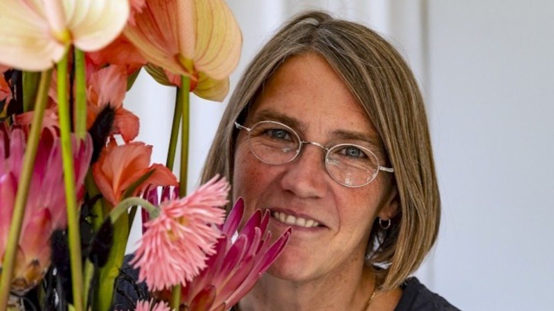 Eine Frau mit Brille l&auml;chelt neben einem farbenfrohen Blumenarrangement mit rosa und orangefarbenen Bl&uuml;ten., &copy; Mercedes-Benz AG
