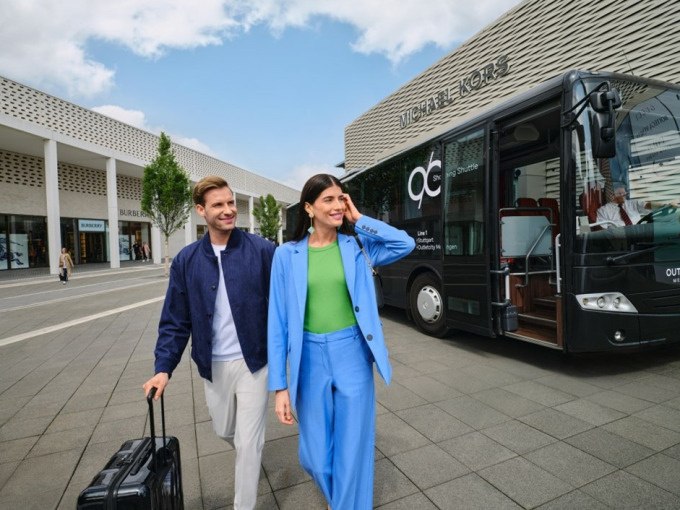 Ein Paar mit Koffern l&auml;uft vor einem Bus in der Outletcity Metzingen. Im Hintergrund sind Gesch&auml;fte und ein Michael Kors Laden zu sehen., &copy; Schlienz-Tours GmbH &amp; Co.KG