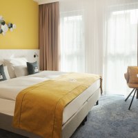Modernes Hotelzimmer mit gelber Wand, Doppelbett, dekorativen Kissen, kleinem Tisch, Stuhl und Wand-TV. Helle Vorhänge lassen Licht herein., © the niu Kettle - Novum Hospitality Modernes Hotelzimmer mit gelber Wand, Doppelbett, dekorativen Kissen, kleinem Tisch, Stuhl und Wand-TV. Helle Vorhänge lassen Licht herein., © the niu Kettle - Novum Hospitality