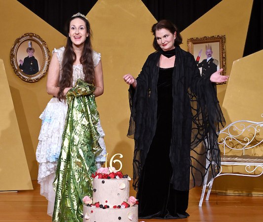 Zwei Frauen in einem M&auml;rchenmusical, eine in einem wei&szlig;en Kleid mit Krone, die andere in einem schwarzen Umhang, stehen hinter einer gro&szlig;en Torte., &copy; Stadtverwaltung Kirchheim unter Teck - Kulturring