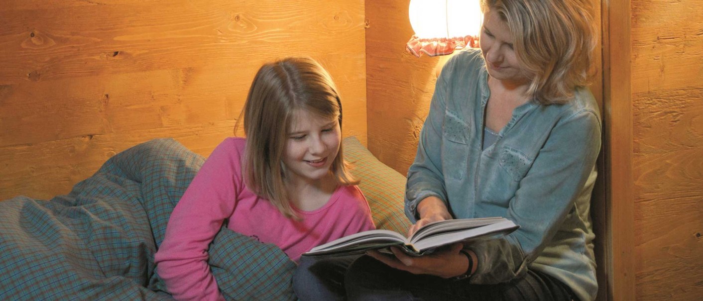 Eine Frau liest einem Mädchen in einem gemütlichen Holzraum ein Buch vor. Beide wirken entspannt und lächeln., © Tripsdrill Natur-Resort GmbH