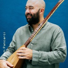 Ein Mann mit Bart hält eine Saz und lächelt vor einem blauen Hintergrund., © links im Bild Ein Mann mit Bart hält eine Saz und lächelt vor einem blauen Hintergrund., © links im Bild