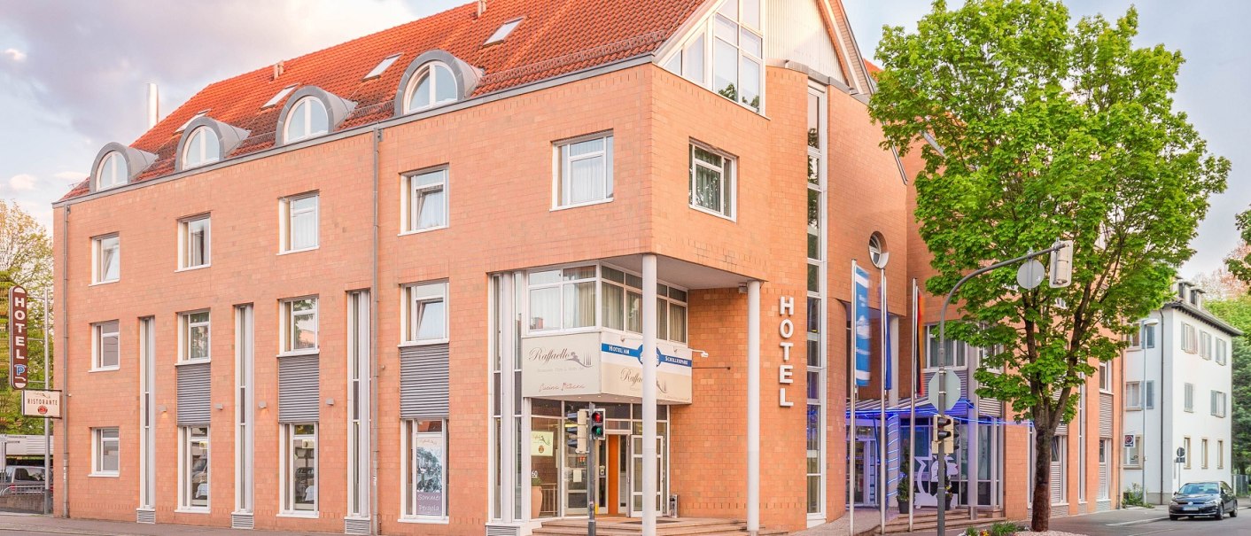 Modernes Hotelgebäude mit rotem Ziegeldach und großen Fenstern an einer Straßenecke, umgeben von Bäumen., © TOMAS Modernes Hotelgebäude mit rotem Ziegeldach und großen Fenstern an einer Straßenecke, umgeben von Bäumen., © TOMAS