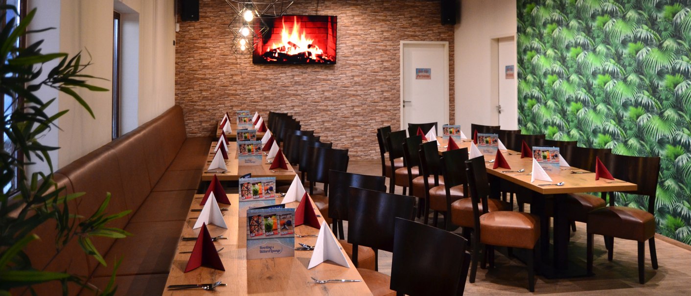 Ein langer Tisch in einer Lounge, dekoriert mit roten und weißen Servietten. An der Wand ein Bildschirm mit Kaminfeuer, daneben eine grüne Tapete., © Bowling & Billard Lounge