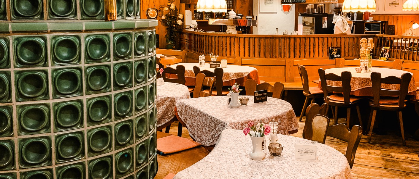 Gemütliches Restaurant mit Holzmöbeln, Tischdecken und einem grünen Kachelofen. Lampen hängen von der Decke, Bar im Hintergrund., © Weinstube Kachelofen