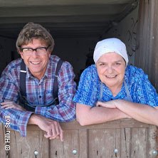 Zwei Personen in ländlicher Kleidung lehnen sich lächelnd über eine Holztür in einem rustikalen Gebäude., © links im Bild