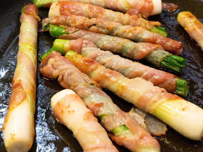Lauchstangen, umwickelt mit Speck, braten in einer Pfanne. Die goldbraune Kruste deutet auf knusprige Textur hin., © Kultweine-Kochwelten-Kunstücke - Kochschule Sonja Lenz Lauchstangen, umwickelt mit Speck, braten in einer Pfanne. Die goldbraune Kruste deutet auf knusprige Textur hin., © Kultweine-Kochwelten-Kunstücke - Kochschule Sonja Lenz