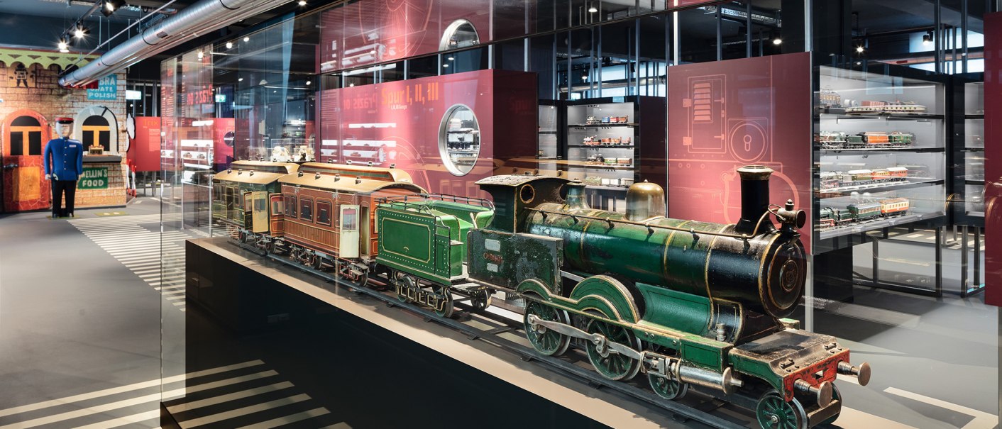 Eine grüne Modelllokomotive in einer Glasvitrine, umgeben von weiteren Modelleisenbahnen in einem Museumsausstellungsraum., © Märklineum