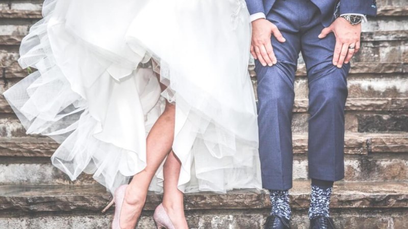 Ein Brautpaar sitzt auf einer Steintreppe. Die Braut tr&auml;gt ein wei&szlig;es Kleid und glitzernde High Heels, der Br&auml;utigam einen blauen Anzug mit gemusterten Socken., &copy; Canva