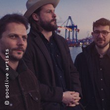 Drei Männer stehen vor einem Hafen bei Dämmerung. Im Hintergrund sind Kräne zu sehen. Sie tragen Jacken und einer hat einen Hut auf., © links im Bild