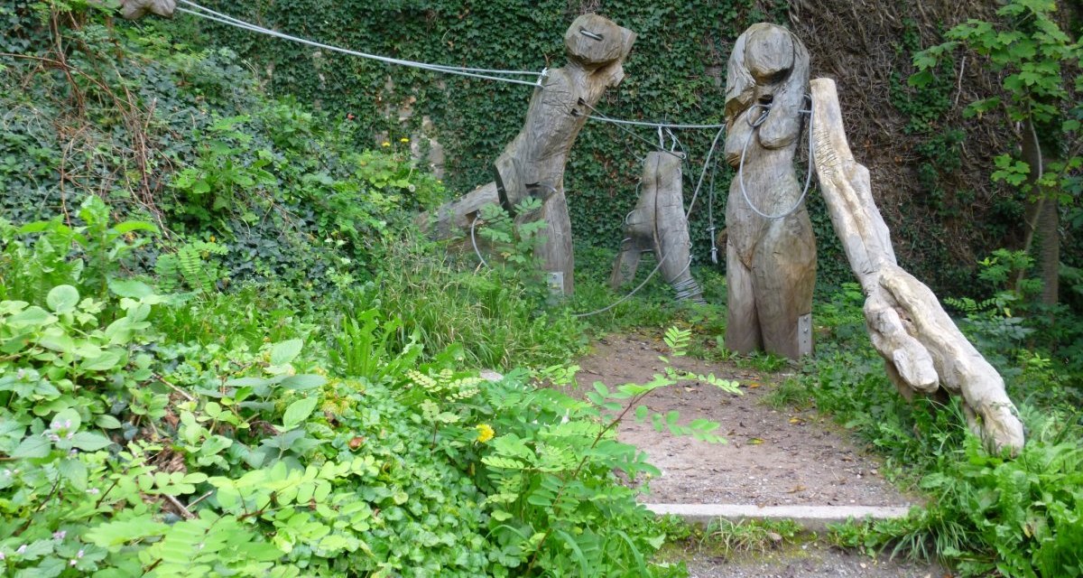 Holzskulpturen auf einem Pfad, umgeben von &uuml;ppigem Gr&uuml;n und Kletterpflanzen. Die Figuren sind durch Seile verbunden., &copy; Natur.Nah. Sch&ouml;nbuch & Heckeng&auml;u