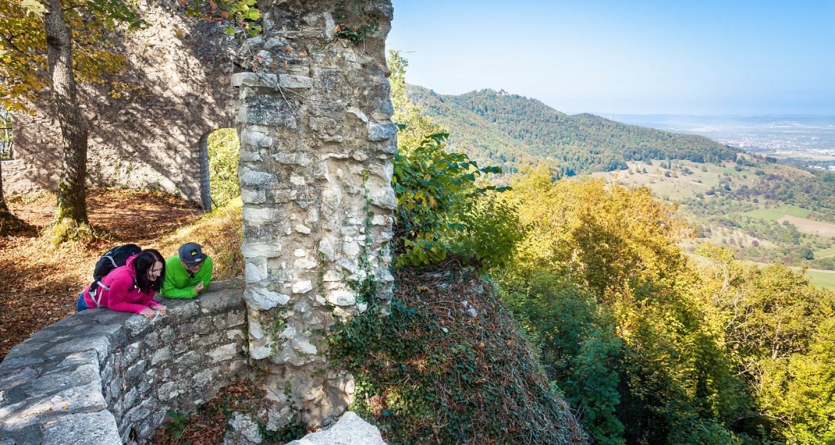 Zwei Personen in bunter Kleidung betrachten eine alte Steinmauer in einer bewaldeten Landschaft mit weitem Ausblick., © hochgehberge Zwei Personen in bunter Kleidung betrachten eine alte Steinmauer in einer bewaldeten Landschaft mit weitem Ausblick., © hochgehberge