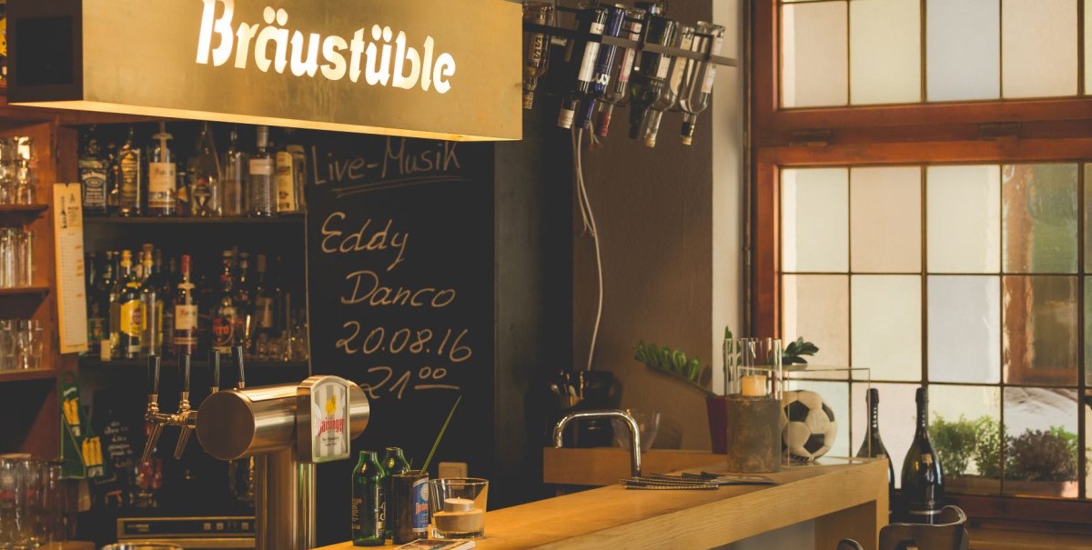 Gemütliche Bar mit Holztheke, Flaschenregal und Schild 'Bräustüble'. Schwarzes Brett kündigt Live-Musik an. Fenster im Hintergrund.