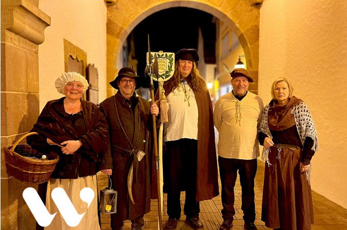 Fünf Personen in historischen Kostümen stehen unter einem beleuchteten Torbogen. Sie tragen Hüte, Umhänge und halten Laternen und Stäbe., © Stadt Winnenden