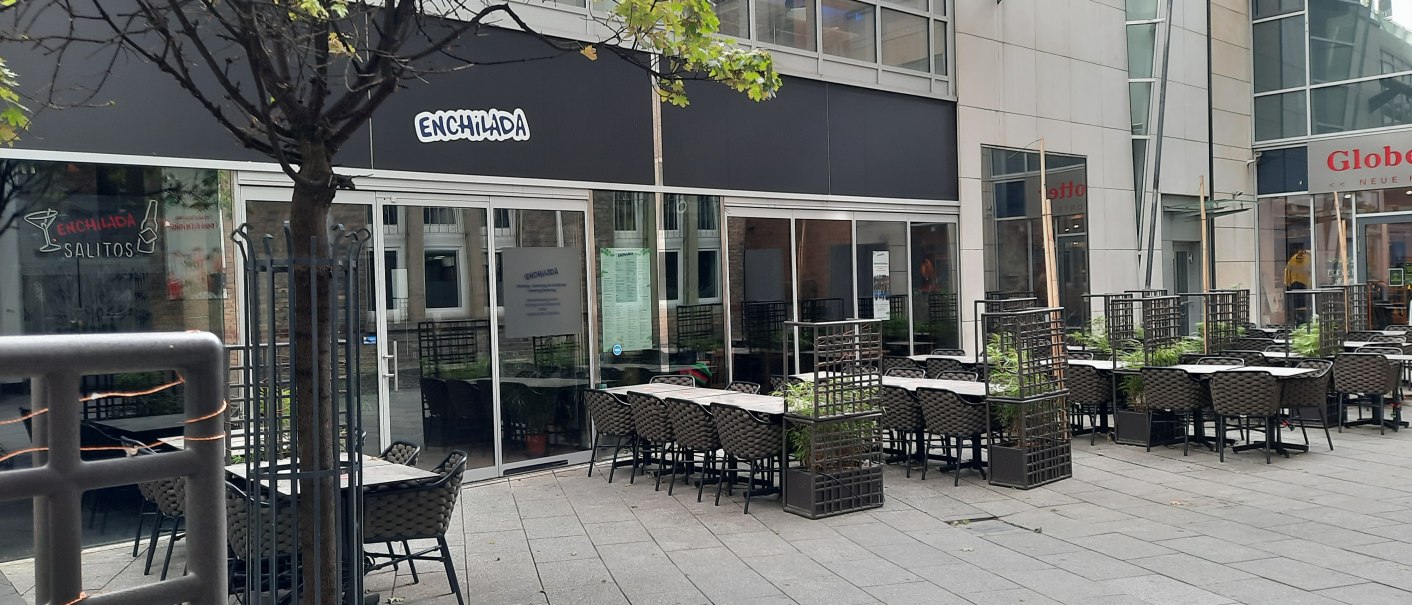 Außenbereich eines Restaurants namens Enchilada mit leeren Tischen und Stühlen. Ein Baum und Pflanzen trennen die Sitzbereiche., © SMG Außenbereich eines Restaurants namens Enchilada mit leeren Tischen und Stühlen. Ein Baum und Pflanzen trennen die Sitzbereiche., © SMG