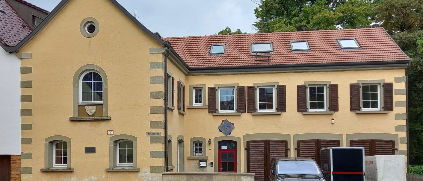 zweist&ouml;ckiges historisches Geb&auml;ude mit gelber Fassade und roten Dachziegeln. Im Untergescho&szlig; auf der rechten Seite drei rotbraune Doppelt&uuml;ren. Vor den T&uuml;ren stehen ein schwarzer PKW und zweiAnh&auml;nger, &copy; Petra Natzkowski