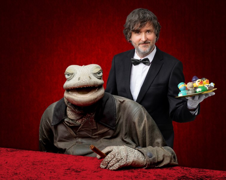 Ein Mann im Anzug mit Fliege hält ein Tablett mit Gummienten. Neben ihm sitzt eine große Echsenpuppe mit Zigarre. Roter Hintergrund., © Scala Kultur Live gGmbH Ein Mann im Anzug mit Fliege hält ein Tablett mit Gummienten. Neben ihm sitzt eine große Echsenpuppe mit Zigarre. Roter Hintergrund., © Scala Kultur Live gGmbH
