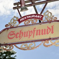 Ein dekoratives Schild mit der Aufschrift 'Schupfnudl' hängt vor einem blauen Himmel. Es gehört zu einem Restaurant., © Schupfnudl