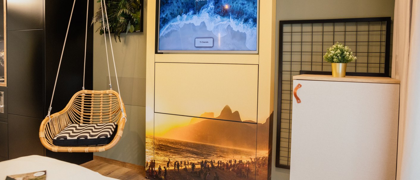 Ein gemütlicher Raum mit einem Hängesessel, einem Fernseher mit Strandbild und einem kleinen Schrank mit Pflanze., © Rioca Stuttgart Posto 4 GmbH