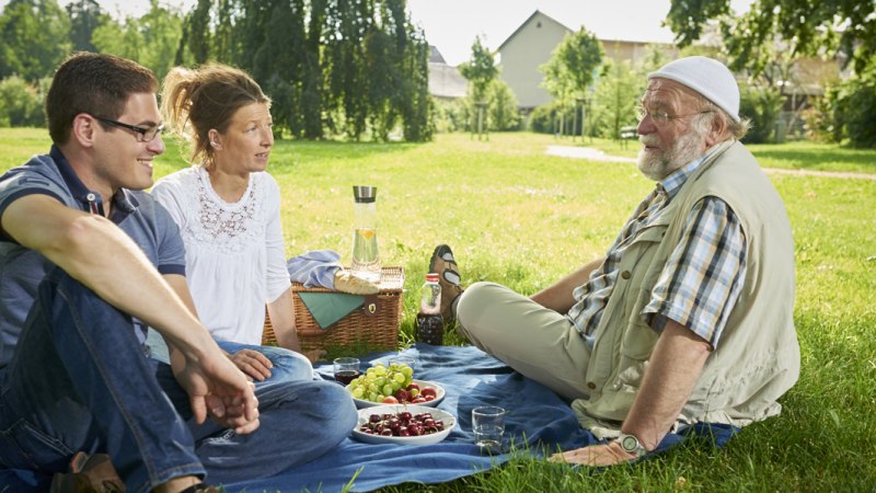 Drei Personen sitzen auf einer Decke im Park und genießen ein Picknick mit Obst und Getränken., © Jean-Claude Winkler Drei Personen sitzen auf einer Decke im Park und genießen ein Picknick mit Obst und Getränken., © Jean-Claude Winkler