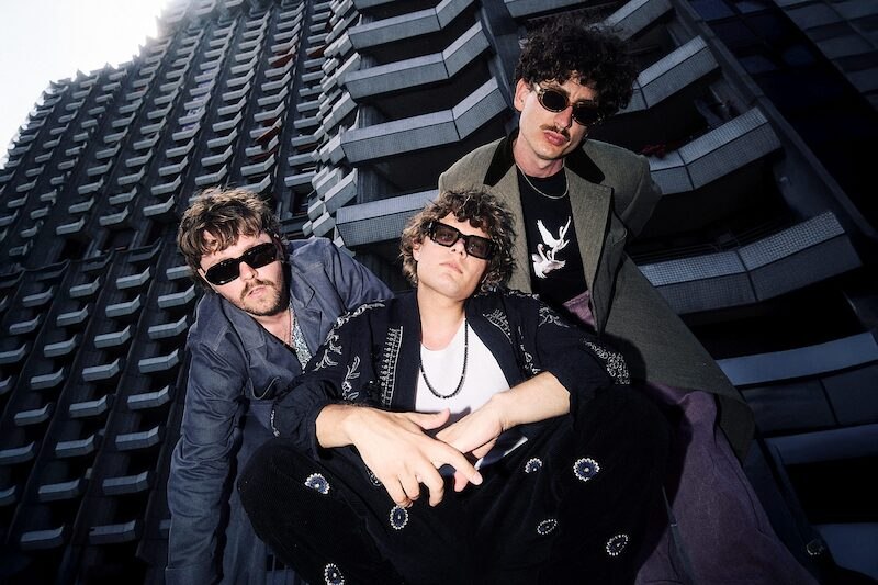 Drei Männer mit Sonnenbrillen posieren lässig vor einem modernen, hohen Gebäude. Sie tragen modische Kleidung und schauen in die Kamera., © Kulturhaus Schwanen Drei Männer mit Sonnenbrillen posieren lässig vor einem modernen, hohen Gebäude. Sie tragen modische Kleidung und schauen in die Kamera., © Kulturhaus Schwanen