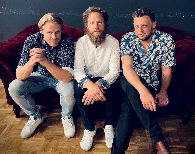 Drei M&auml;nner sitzen auf einem roten Sofa, l&auml;ssig gekleidet in Hemden und Jeans, und blicken in die Kamera., &copy; Foto Daniel Mudrack