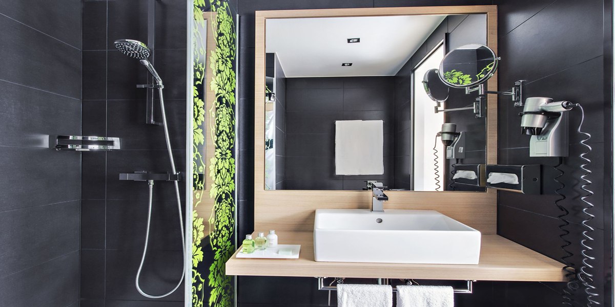 Modernes Badezimmer mit schwarzer Fliesenwand, Waschbecken, großem Spiegel, Dusche und Haartrockner. Grüne Pflanzenmuster als Dekoration., © NH Ludwigsburg