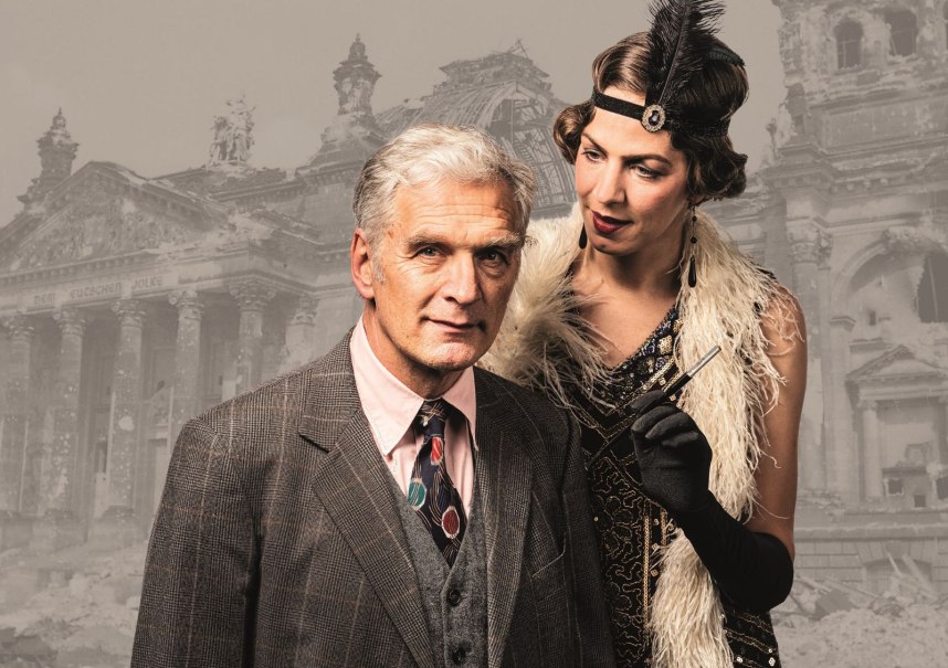 Ein Mann im Anzug und eine Frau im 1920er-Jahre-Kleid stehen vor einem historischen Gebäude. Die Frau trägt eine Feder im Haar.