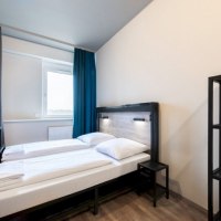 Modernes Doppelzimmer mit zwei Einzelbetten, einem kleinen Nachttisch und einem Regal. Helle Wände und Vorhänge in Blau und Grau., © A&O Hostels Marketing GmbH