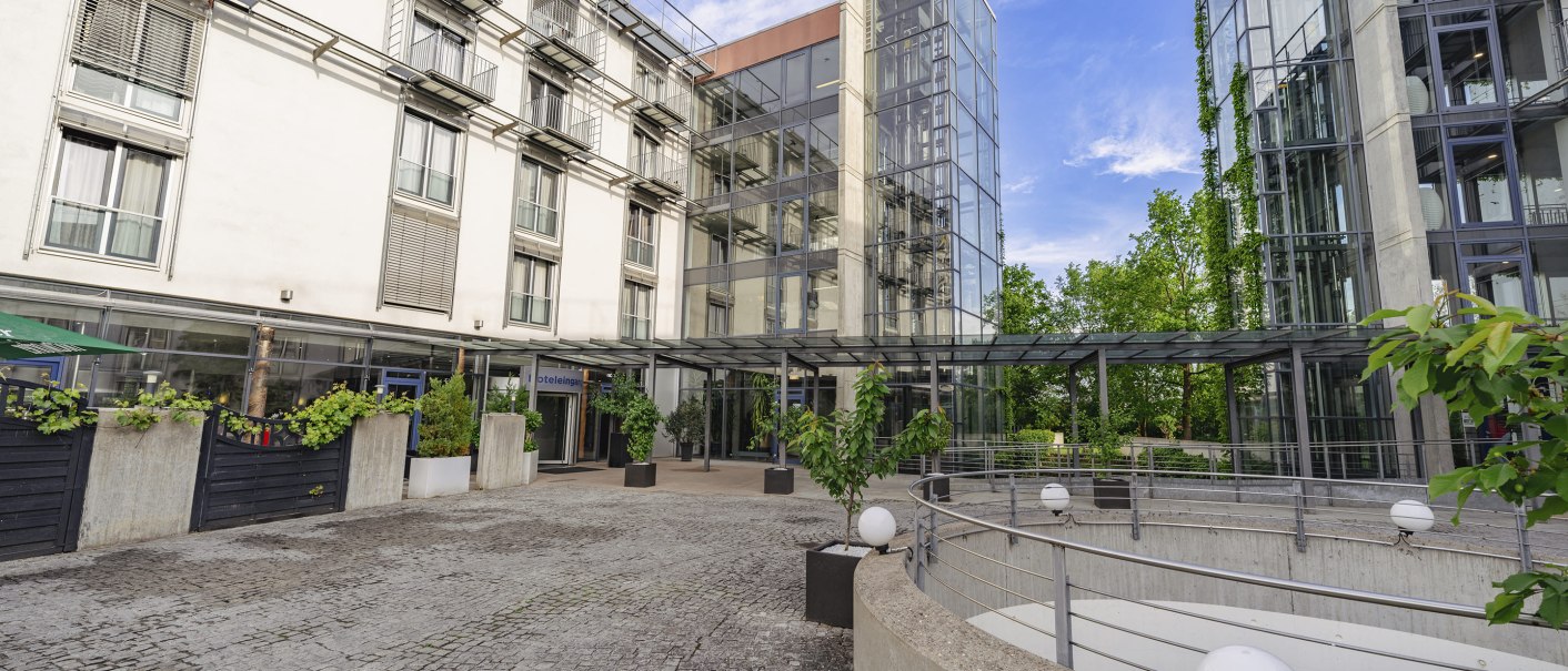 Modernes Gebäude mit Glasfassade, Pflanzen im Eingangsbereich und gepflastertem Vorplatz. Klare Linien und viel Glas prägen das Design., © PLAZA Hotelgroup GmbH Modernes Gebäude mit Glasfassade, Pflanzen im Eingangsbereich und gepflastertem Vorplatz. Klare Linien und viel Glas prägen das Design., © PLAZA Hotelgroup GmbH
