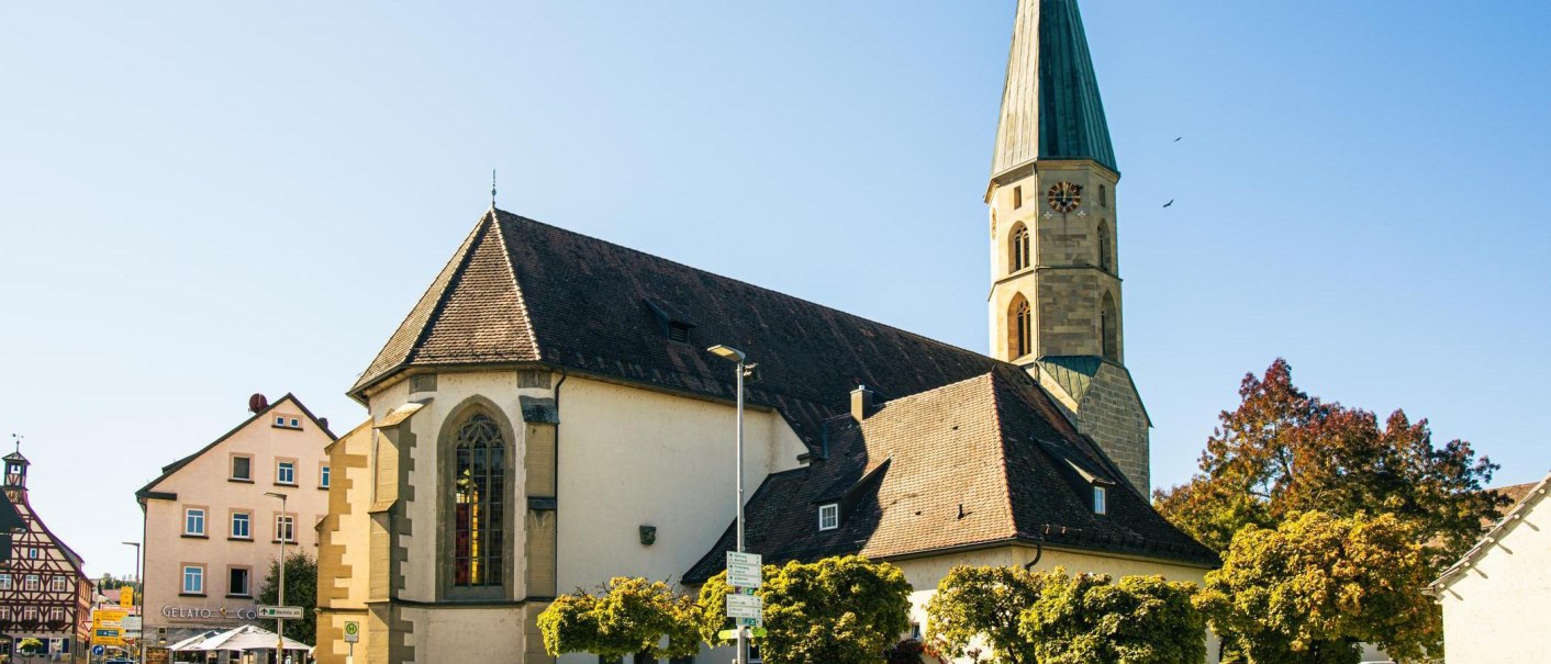 Eine Kirche mit spitzem Turm in Gaildorf, umgeben von Bäumen und einem Fachwerkhaus im Hintergrund bei klarem Himmel., © Stuttgart-Marketing GmbH, Sarah Schmid