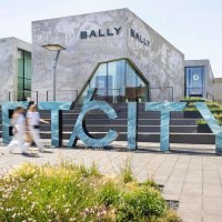 Gro&szlig;e Buchstaben formen das Wort 'OUTLETCITY' vor modernen Geb&auml;uden. Menschen gehen vorbei, w&auml;hrend Blumen im Vordergrund bl&uuml;hen., &copy; OUTLETCITY AG
