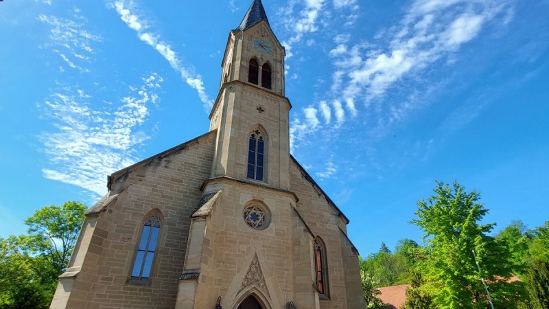 Die Evangelische Schlosskirche Remseck-Hochberg vor blauem Himmel mit dekorativen Wolken. Der Kirchturm ragt in die H&ouml;he, umgeben von B&auml;umen.