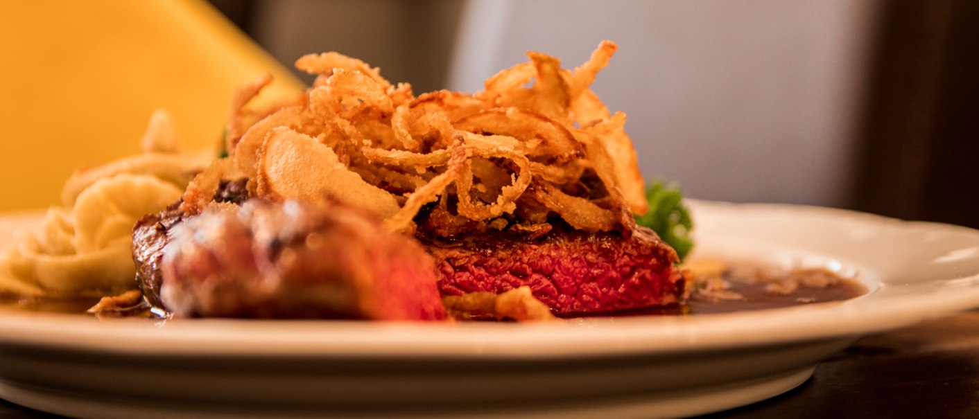Ein saftiges Steak mit knusprigen Röstzwiebeln und Beilage auf einem weißen Teller, appetitlich angerichtet., © Restaurant Café Hirsch Ein saftiges Steak mit knusprigen Röstzwiebeln und Beilage auf einem weißen Teller, appetitlich angerichtet., © Restaurant Café Hirsch