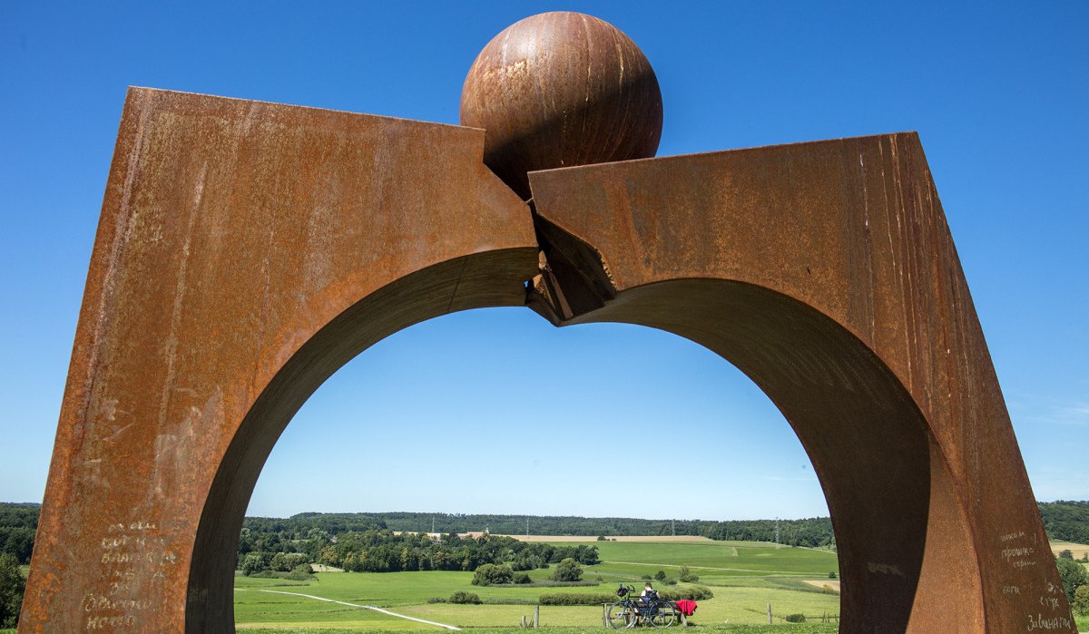 Rostige Metallskulptur mit Kugel auf einem Hügel, darunter grüne Landschaft und blauer Himmel. Ein Fahrradfahrer sitzt im Hintergrund., © Stuttgart Marketing GmbH Rostige Metallskulptur mit Kugel auf einem Hügel, darunter grüne Landschaft und blauer Himmel. Ein Fahrradfahrer sitzt im Hintergrund., © Stuttgart Marketing GmbH