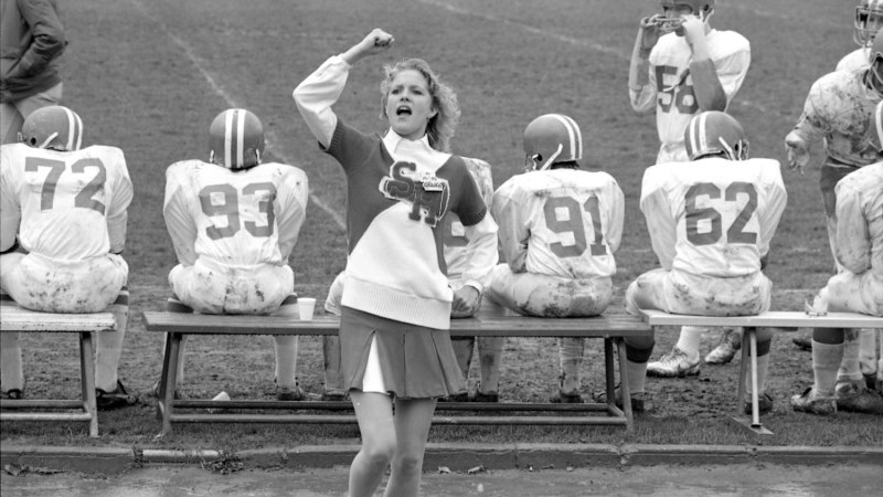 Ein Cheerleader in Uniform feuert ein Football-Team an, das auf einer Bank am Spielfeldrand sitzt. Die Spieler tragen Helme und schmutzige Trikots., &copy; Ludwigsburg Museum