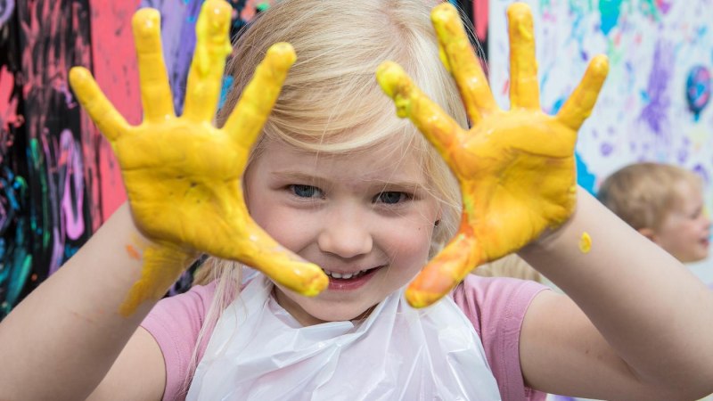 Ein fr&ouml;hliches Kind zeigt seine mit gelber Farbe bemalten H&auml;nde vor einem bunten Hintergrund. Es tr&auml;gt ein wei&szlig;es L&auml;tzchen., &copy; Tourismus & Events Ludwigsburg
