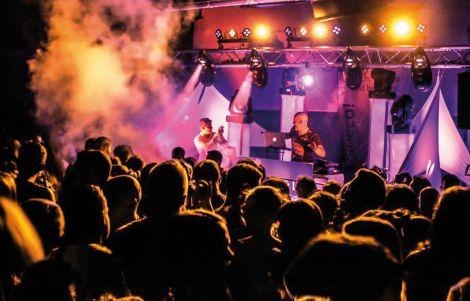 Eine lebhafte Clubszene mit einem DJ auf der B&uuml;hne, umgeben von bunten Lichtern und einer tanzenden Menge., &copy; Esecco.Nacht. Foto: Teamwerk