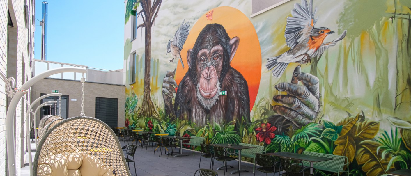 Terrasse mit Hängesesseln und Tischen, Wandgemälde zeigt einen Schimpansen und Vögel vor einem orangefarbenen Hintergrund., © 13 6 Design GmbH (Judenhof 10, 89073 Ulm) Terrasse mit Hängesesseln und Tischen, Wandgemälde zeigt einen Schimpansen und Vögel vor einem orangefarbenen Hintergrund., © 13 6 Design GmbH (Judenhof 10, 89073 Ulm)