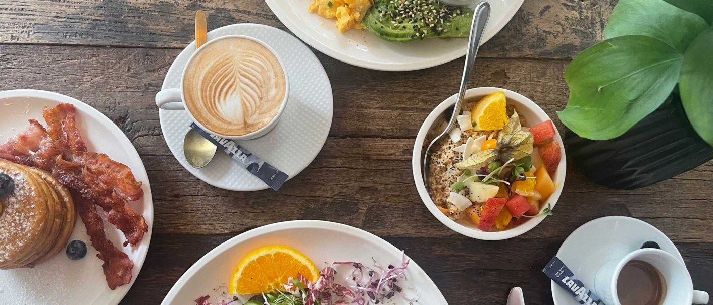 Ein reich gedeckter Frühstückstisch mit Kaffee, Pancakes mit Speck, Avocado mit Rührei und einer Schale mit frischem Obst., © SMG