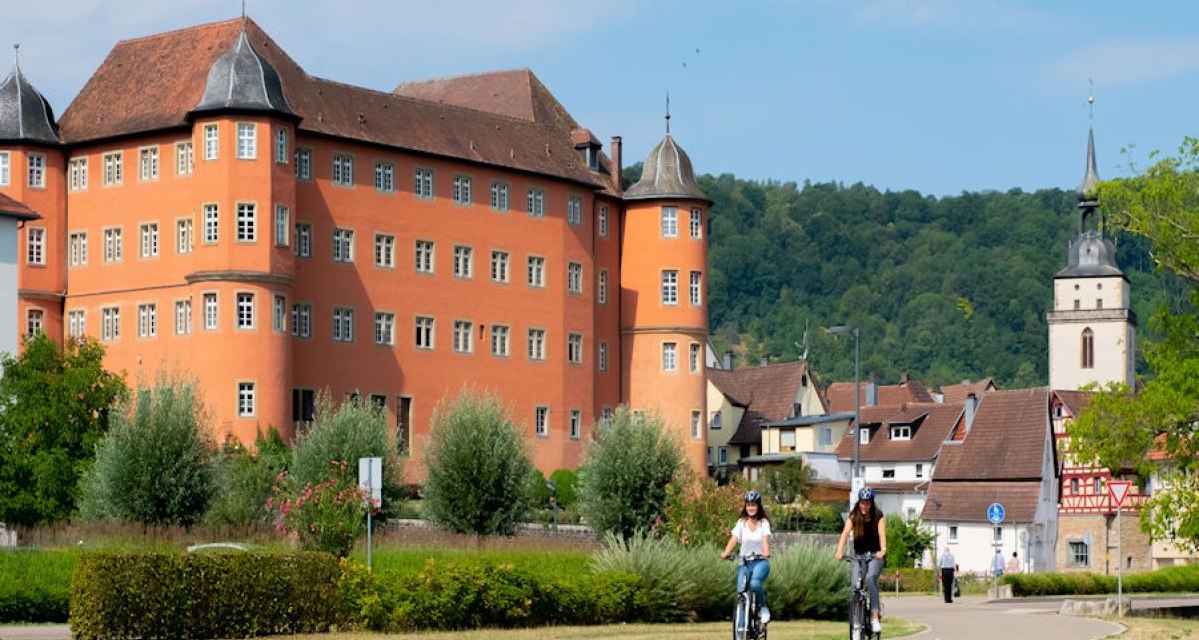 Das Bild zeigt das Schloss in Künzelsau mit seiner markanten roten Fassade. Im Vordergrund fahren zwei Personen auf Fahrrädern. Im Hintergrund ist eine Kirche sichtbar., © Stadt Künzelsau / Olivier Schniepp Das Bild zeigt das Schloss in Künzelsau mit seiner markanten roten Fassade. Im Vordergrund fahren zwei Personen auf Fahrrädern. Im Hintergrund ist eine Kirche sichtbar., © Stadt Künzelsau / Olivier Schniepp
